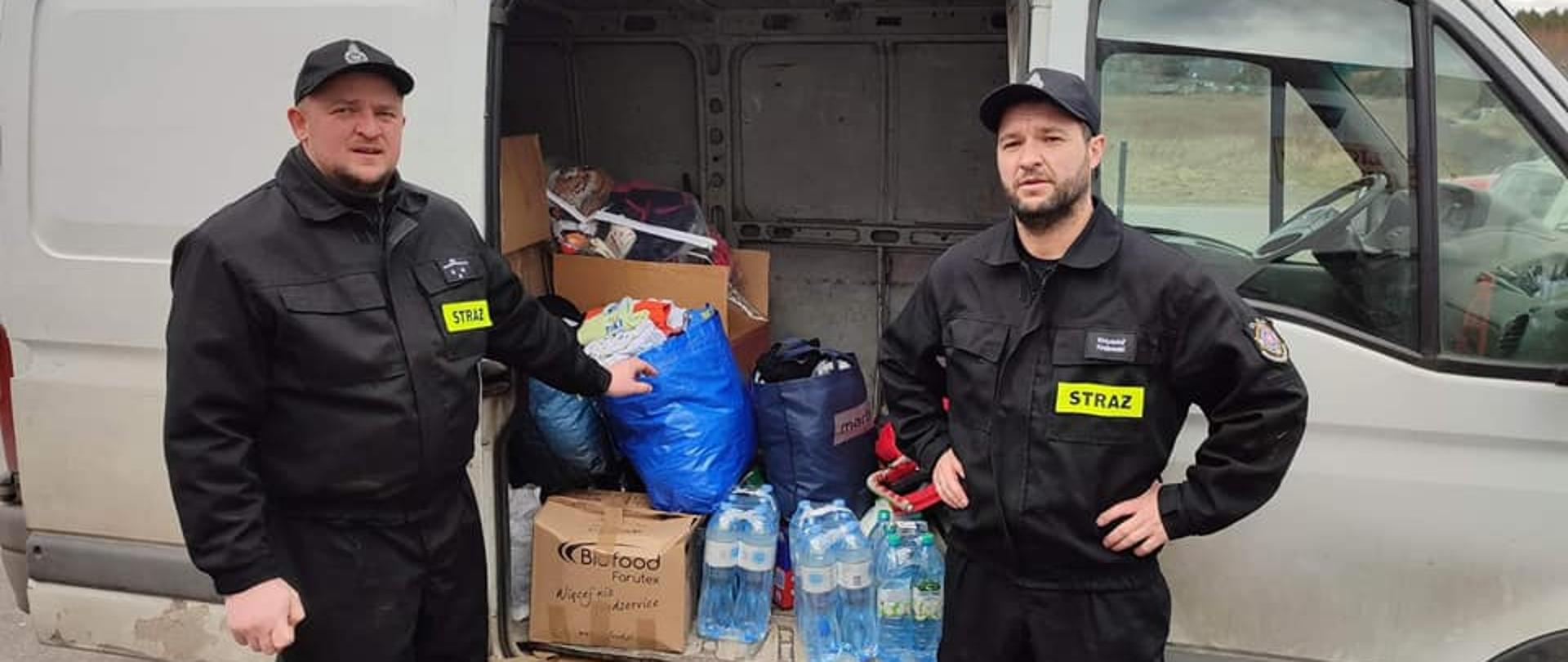 Na pierwszym planie dwaj strażacy w czarnych ubraniach koszarowych, stojący przodem do osoby robiącej zdjęcie, za strażakami widać prawy bok białego busa, który ma otwarte boczne drzwi od przedziału bagażowego. W przedziale bagażowym są zebrane dary w kartonach, torbach foliowych. 