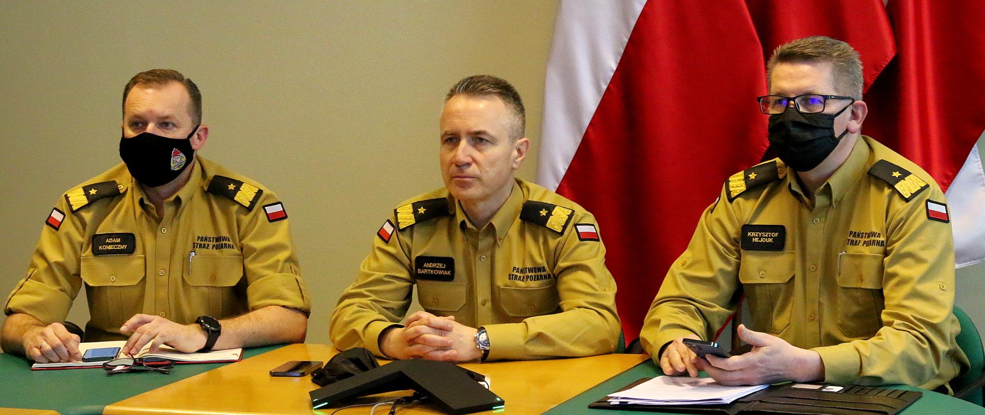 Po lewej stronie zastępca komendanta głównego PSP nadbryg. Adam Konieczny, następnie komendant główny PSP, nadbryg. Andrzej Bartkowiak, po prawej stronie zastępca KG PSP nadbryg. Krzysztof Hejduk. Wszyscy trzej siedzą przy stole podczas wideokonferencji, ubrani są w nowe umundurowanie służbowe koloru "musztardowego"