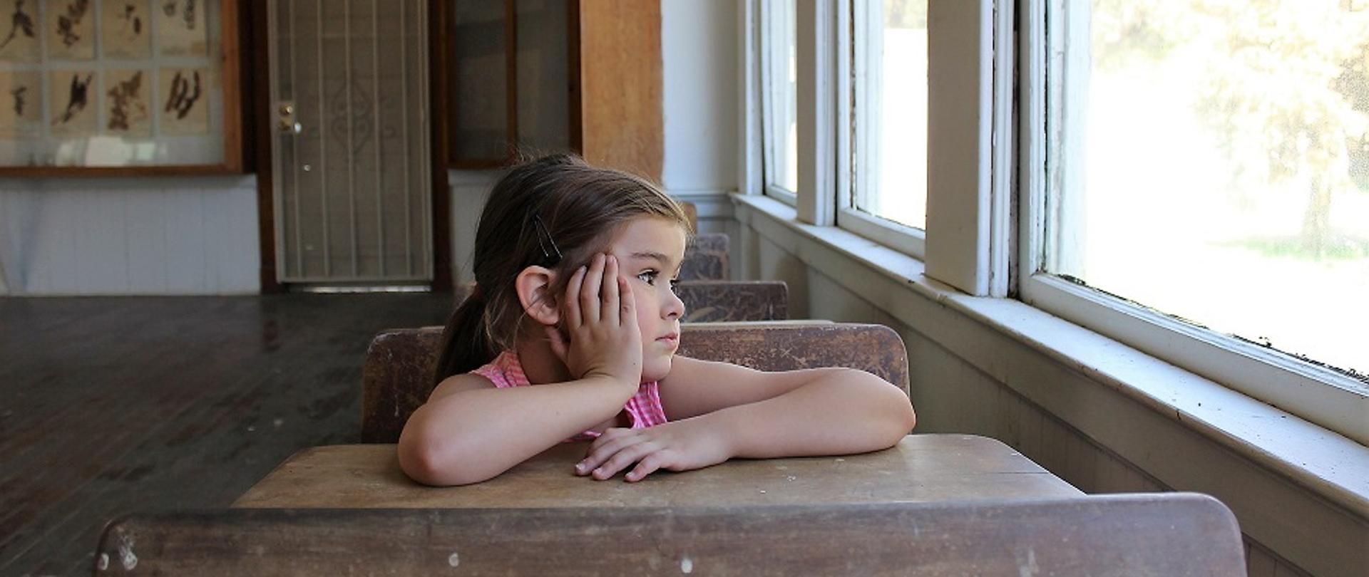 dziewczynka siedząca sama w ławce szkolnej w pustej klasie, podpierająca głowę ręką, patrząca przez zamknięte okna