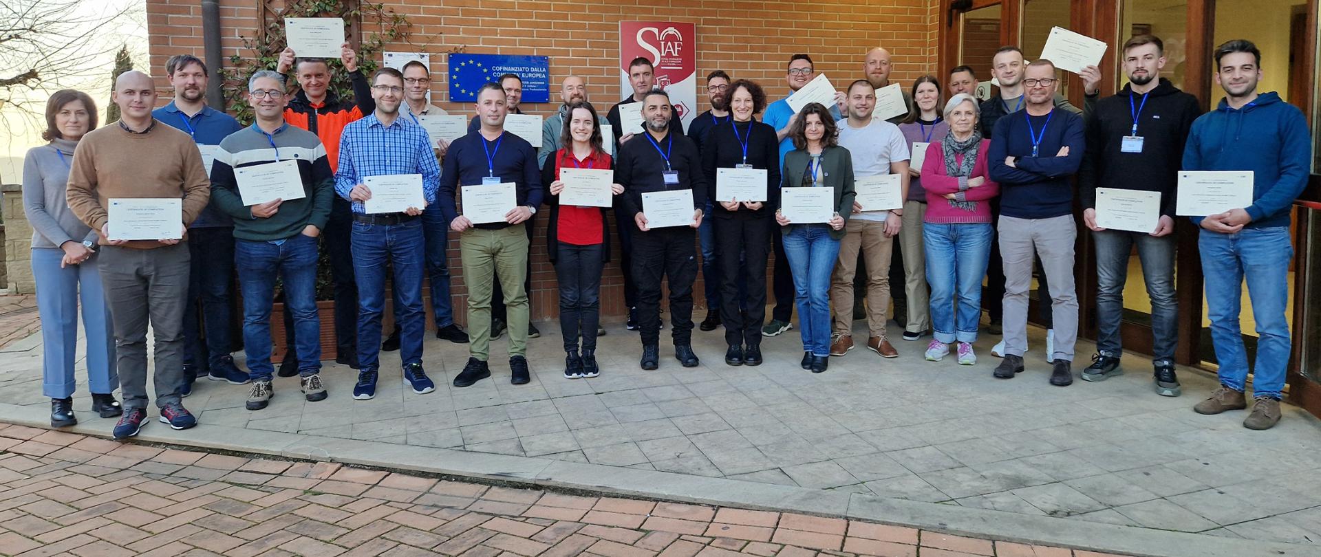 Zdjęcie pamiątkowe uczestników szkolenia. Grupa ludzi przed budynkiem z cegły. Każdy z nich trzyma w ręku certyfikat. 