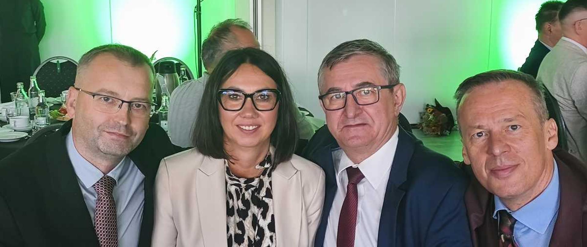 Opis ogólnyZdjęcie przedstawia cztery osoby – kobietę i trzech mężczyzn – siedzące blisko siebie w rzędzie i uśmiechające się do obiektywu. Wszyscy są elegancko ubrani, co sugeruje oficjalne lub uroczyste wydarzenie. Kadrowanie obejmuje postaci od pasa w górę. W tle widoczna jest sala bankietowa z charakterystycznym, zielonym podświetleniem.Szczegóły postaci (od lewej do prawej)Pierwszy mężczyzna (po lewej): Ma krótkie, ciemne, lekko szpakowate włosy oraz zarost. Nosi okulary w prostokątnych oprawkach. Ubrany jest w czarną marynarkę, jasnoniebieską koszulę oraz wzorzysty krawat w drobną kratkę w odcieniach czerwieni i bieli. Jest lekko pochylony w stronę siedzącej obok kobiety.Kobieta (druga od lewej): Ma ciemne włosy sięgające do ramion, obcięte na prosto z przedziałkiem pośrodku. Nosi duże okulary w wyrazistych, czarnych oprawkach. Ma na sobie elegancką bluzkę w czarno-białe cętki (wzór panterki) oraz jasnobeżową marynarkę.Drugi mężczyzna (trzeci od lewej): To starszy mężczyzna o krótkich, siwiejących włosach, również noszący okulary. Jest ubrany w ciemnoniebieski (granatowy) garnitur, białą koszulę i gładki, bordowy krawat. Z kieszonki jego marynarki (brustaszy) wystaje róg białej poszetki.Trzeci mężczyzna (po prawej): Ma krótkie, ciemne włosy zaczesane do góry. Ubrany jest w ciemnobordową (lub ciemnobrązową) marynarkę, błękitną koszulę oraz wzorzysty, ciemny krawat. W jego poszetce również znajduje się biała, złożona chusteczka. Podobnie jak pierwszy mężczyzna, pochyla się mocno do środka kadru.Tło i otoczenieGrupa siedzi przy stole, na którym widoczne są elementy zastawy, m.in. szklane butelki i filiżanki. Za nimi, po lewej stronie, stoi w głębi inna kobieta w ciemnym stroju, a po prawej widać fragmenty sylwetek innych gości. Ściany w tle są jasne, ale całe pomieszczenie jest oświetlone intensywnym, neonowozielonym światłem punktowym, które tworzy jaskrawe plamy za głowami fotografowanych osób. Po lewej stronie w tle widoczny jest także czarny głośnik na statywie.