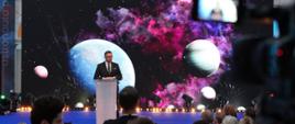 Widok od tyłu na salę, na podwyższeniu za mównicą stoi minister Kulasek i mówi do mikrofonu, za nim wielki ekran z obrazem kilku planet na czarno-fioletowym tle.