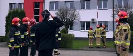 Zdjęcie przedstawia zmianę służbową strażaków podczas podnoszenia flagi państwowej na maszt. Na czele szeregu na pierwszym planie stoi oficer w mundurze wyjściowym z czapką rogatywką i salutuje. Strażacy stoją frontem do siebie w szeregu sześciu na lewej stronie i ośmiu po prawej stronie, wszyscy spoglądają na maszt, na który jest wciągana flaga państwowa przez trzech ratowników. Jeden strażak salutuje podnoszonej na maszcie fladze, po jego lewej stronie strażak stoi w pozycji zasadniczej, po prawo ratownik wciąga flagę, flaga znajduje się na samym szczycie masztu. Wszyscy strażacy są ubrani w ubrania bojowe typu Moratex, na głowie maja heł, na twarzy maseczki ochronne, na nogach kalosze. Ratownicy po lewo ubrani są w ubrania barwy czarnej z odblaskowymi pasami barwy szarej i żółtej a ratownicy po prawej stronie kadru w ubrania barwy jasno brązowej z odblaskowymi pasami barwy szarej i żółtej. W tle żywopłot oraz budynek Komendy Powiatowej w Łowiczu oraz okna należące do części administracyjnej komendy.