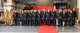 Na zdjęciu grupa funkcjonariuszy Państwowej Straży Pożarnej oraz zaproszonych gości pozuje do wspólnego zdjęcia pamiątkowego podczas uroczystości w hali strażackiej. W tle widoczne są wozy strażackie, czerwony baner z logo PSP oraz flagi Polski i Unii Europejskiej. Część osób ubrana jest w mundury galowe, inni w mundury bojowe lub cywilne garnitury. Na środku znajduje się czerwony dywan, podkreślający rangę wydarzenia.