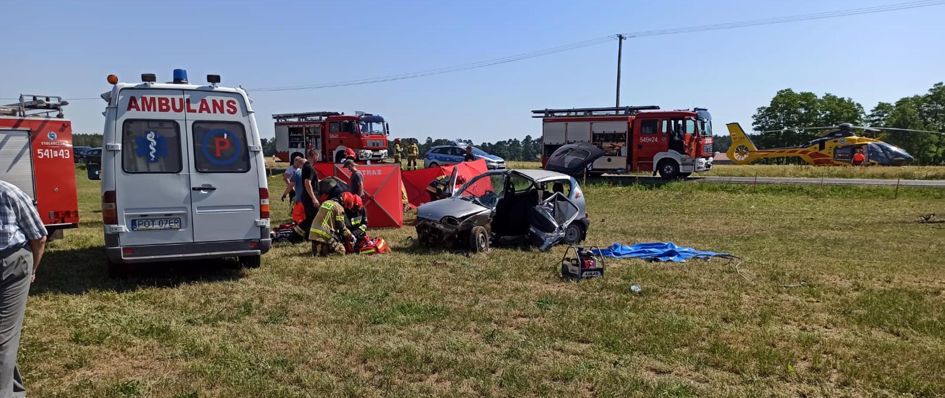 Zdjęcie przedstawia akcję ratowniczą podczas zdarzenia drogowego ze skutkiem śmiertelnym w miejscowości Torzeniec. Na zdjęciu widać wozy ratowniczo - gaśnicze, karetkę, rozbite pojazdy które brały udział w wypadku oraz strażaków podczas działań.