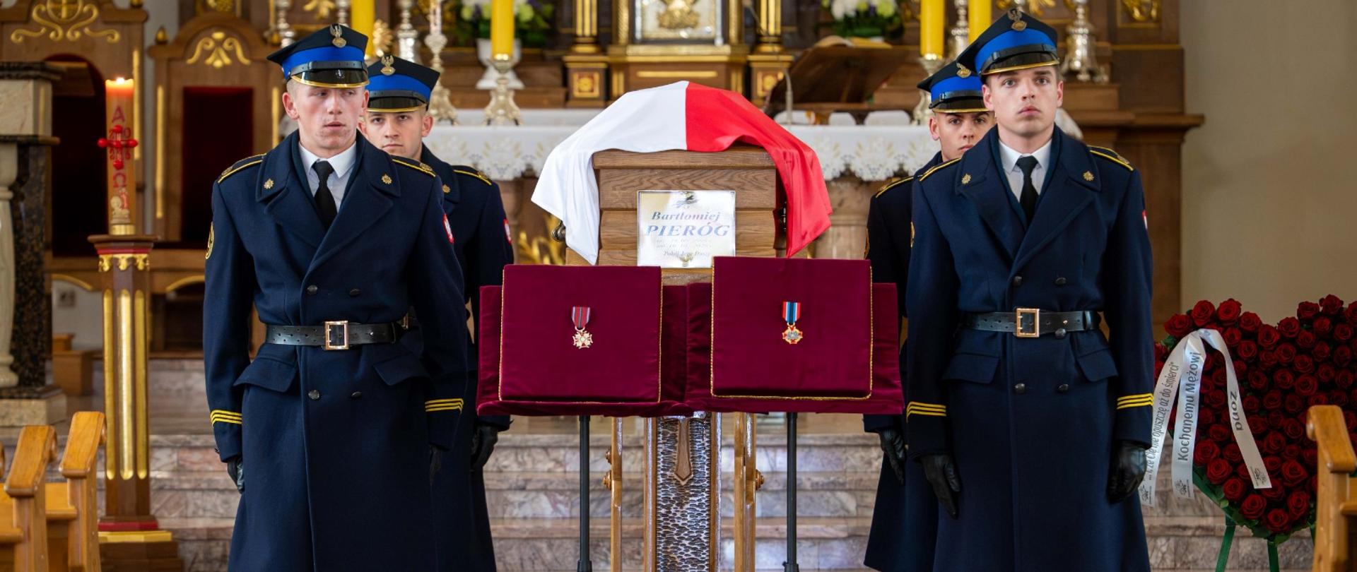 Wnętrze kościoła podczas uroczystości pogrzebowej. Drewniana trumna, przykryta biało-czerwoną flagą Polski, ustawiona jest na katafalku z tabliczką z imieniem i nazwiskiem śp. Bartłomieja Pieroga. Przed trumną, na stojakach, wyeksponowano dwa odznaczenia na poduszkach wyściełanych czerwonym aksamitem. Przy katafalku wartę honorową pełnią czterej strażacy w mundurach galowych
