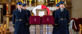 Wnętrze kościoła podczas uroczystości pogrzebowej. Drewniana trumna, przykryta biało-czerwoną flagą Polski, ustawiona jest na katafalku z tabliczką z imieniem i nazwiskiem śp. Bartłomieja Pieroga. Przed trumną, na stojakach, wyeksponowano dwa odznaczenia na poduszkach wyściełanych czerwonym aksamitem. Przy katafalku wartę honorową pełnią czterej strażacy w mundurach galowych