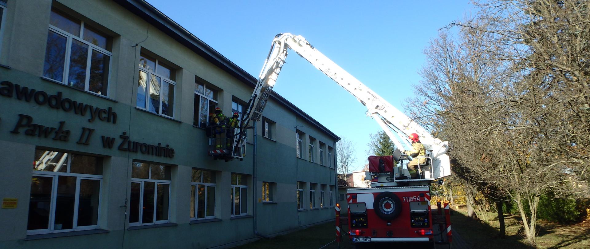 Przed budynkiem szkoły stoi rozstawiony samochód ratowniczo- gaśniczy- podnośnik, którym operuje jeden ze strażaków. Kosz podnośnika podniesiony jest na wysokości okna na piętrze budynku. W koszu znajduje się dwóch strażaków w umundurowaniu specjalnym.
