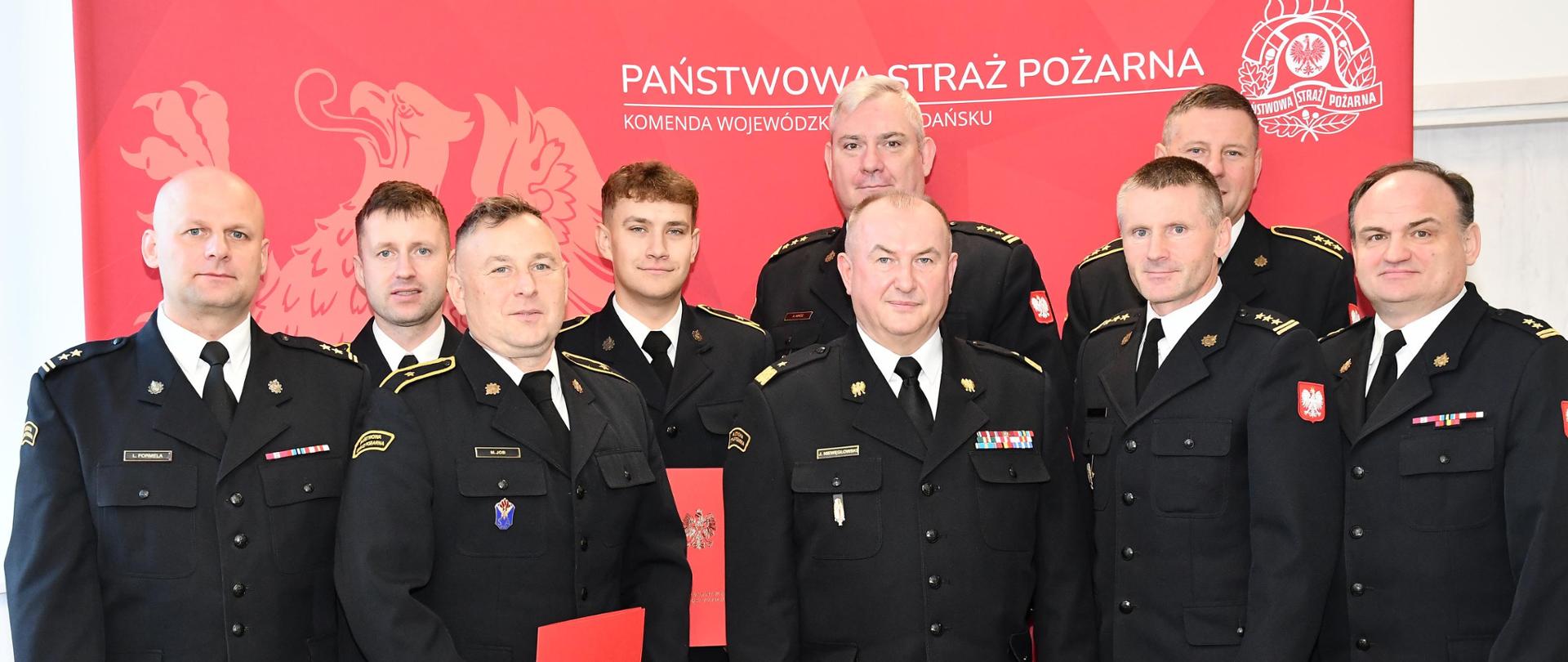 9 strażaków w mundurach wyjściowych stoi do zdjęcia, część z nich teczki w rękach.