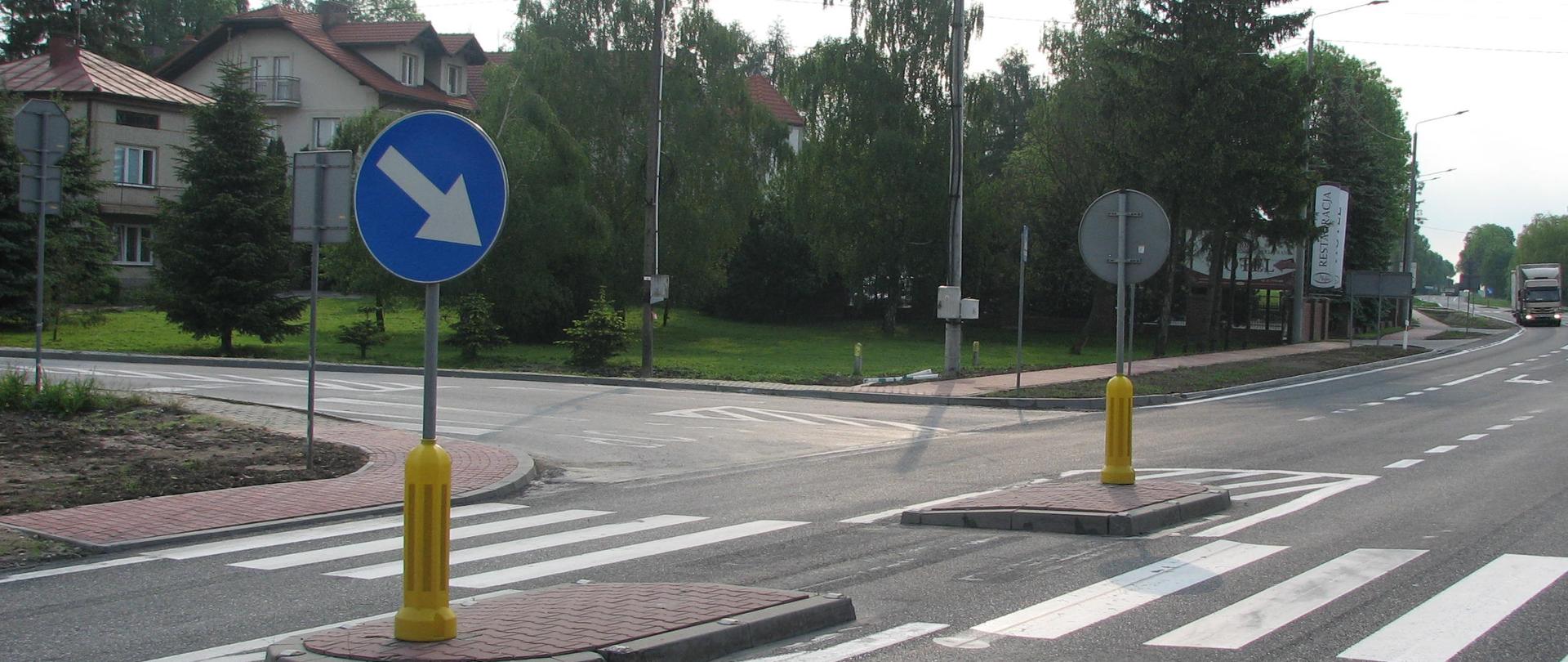DK73 Stopnica - skrzyżowanie z namalowanym przejściem dla pieszych, oznakowany azyl dla pieszych z wysepkami, przy skrzyżowaniu drzewa, dom. W oddali na drodze samochód ciężarowy 