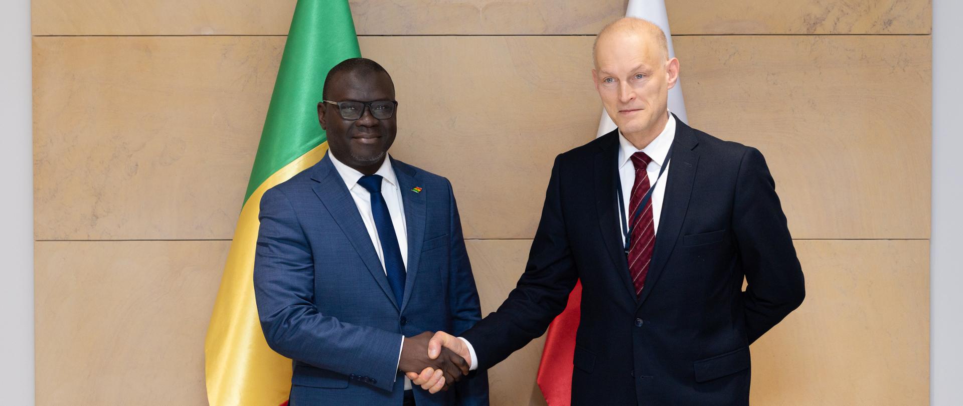 Na zdjęciu dwóch mężczyzn z garniturach w uścisku dłoni. W tle 2 flagi: Polski i Senegalu.