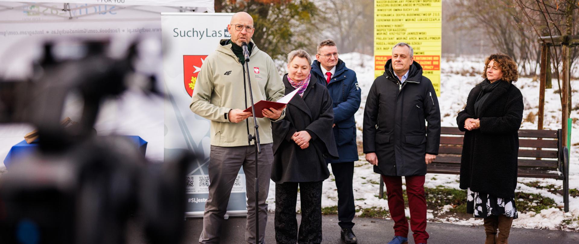 Konferencja prasowa ministra sportu i turystyki Jakuba Rutnickiego na świeżym powietrzu. Pogoda zimowa. Minister mówi do mikrofonu. Za nim, z prawej strony zdjęcia pozostali uczestnicy konferencji (cztery osoby: dwie kobiety i dwóch mężczyzn). Z lewej strony zdjęcia za ministrem namiot z herbem i napisem "Suchy Las". Na pierwszym planie z lewej strony widać niewyraźnie kamerę.