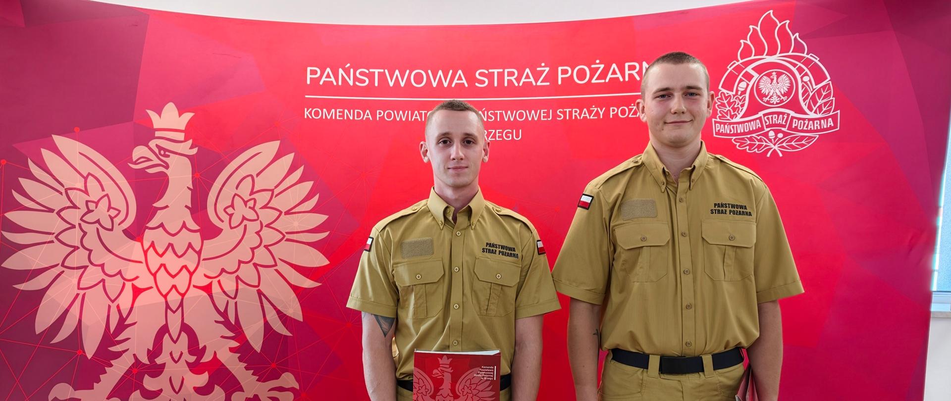 Dwóch nowo przyjętych strażaków na tle czerwonego banneru Państwowej Straży Pożarnej.