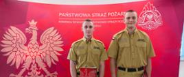 Dwóch nowo przyjętych strażaków na tle czerwonego banneru Państwowej Straży Pożarnej.