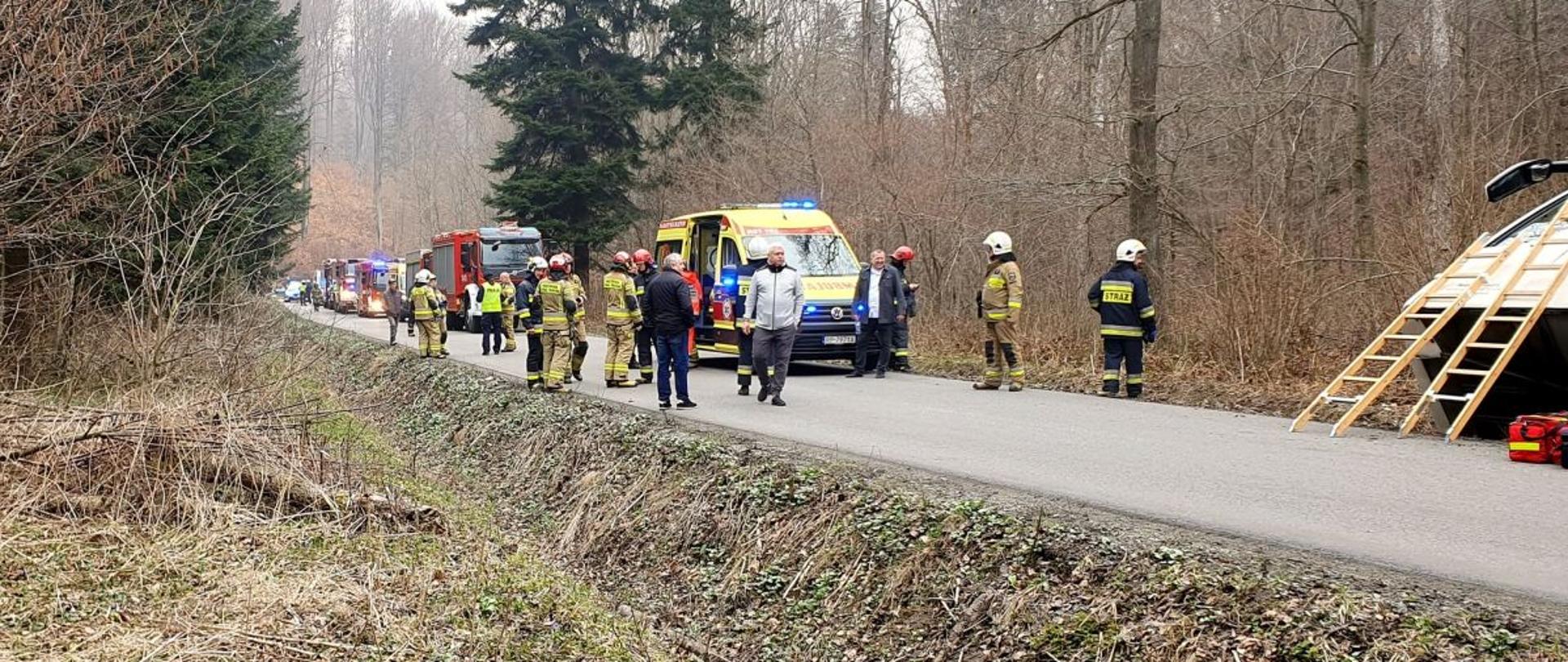 Na zdjęciu znajduje się fragment autobusu który jest przechylony na prawy bok z drogi. Przy nim z przodu oparte są dwa przęsła drabiny nasadkowej i na drodze leżą dwie torby medyczne. Z przodu widać samochody straży pożarnej i strażaków a także ratowników medycznych.