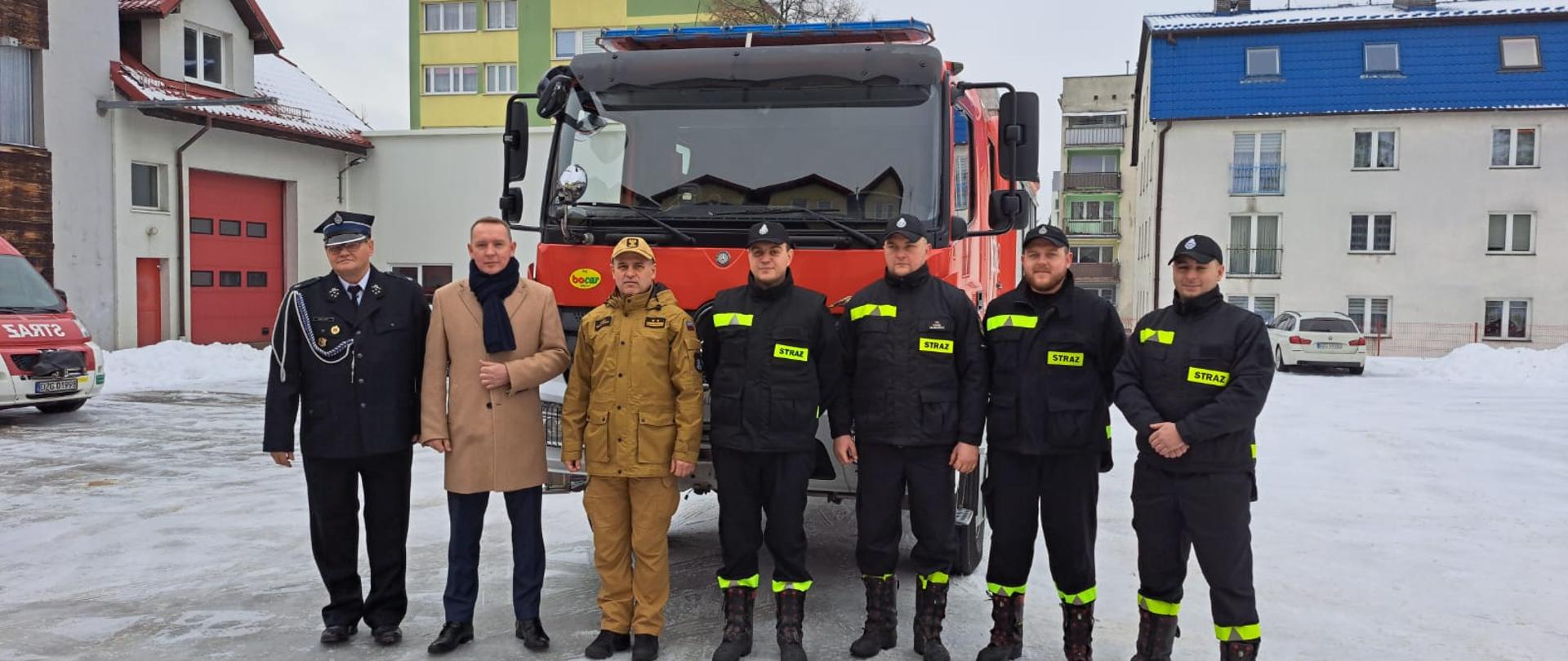 Dzień. Przed samochodem ratowniczo-gaśniczym pozuje do zdjęcia grupa ludzi. Część z nich ubrana jest w ubranie koszarowe w kolorze czarnym i czapki z daszkiem, jedne druh ubrany w mundur wyjściowy i rogatywkę, jedn funkcjonariusz PSP w ubraniu służbowym w kolorze piaskowym oraz czapkę z daszkiem i mężczyzna w płaszczu. W tle budynki mieszkalne.