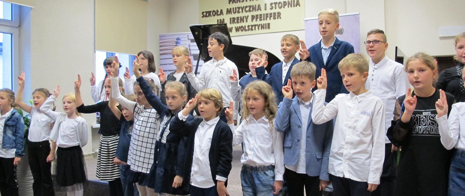 Uczniowie klas pierwszych stoją na scenie w dwóch rzędach. Dzieci podnoszą w górę prawe ręce i składają uroczyste ślubowanie.