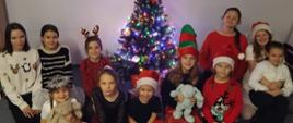 Grupa dzieci pozuje do wspólnego zdjęcia przy udekorowanej choince w sali szkolnej. Choinka stoi pośrodku, ozdobiona kolorowymi lampkami i gwiazdą na czubku. Dzieci siedzą i klęczą wokół niej, ubrane w świąteczne stroje w czerwonych, białych i czarnych kolorach, część ma czapki Mikołaja, opaski z rogami renifera lub świąteczne dodatki. Kilkoro dzieci trzyma pluszowe maskotki. Wszyscy uśmiechają się do aparatu, a scena ma radosny, bożonarodzeniowy charakter.