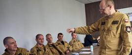 Na zdjęciu widać salę szkoleniową. Przy długim stole siedzi kilku strażaków Państwowej Straży Pożarnej w jednakowych, piaskowych mundurach. Słuchają i patrzą na stojącego przed nimi instruktora, który trzyma w dłoni mały przedmiot i coś tłumaczy. Na stole leżą kartki, długopisy i drobny sprzęt. 