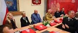 Za stołem siedzą zaproszeni goście, na stole znajdują się torby z logiem PSP z tyłu godło Polski oraz sztandar PSP