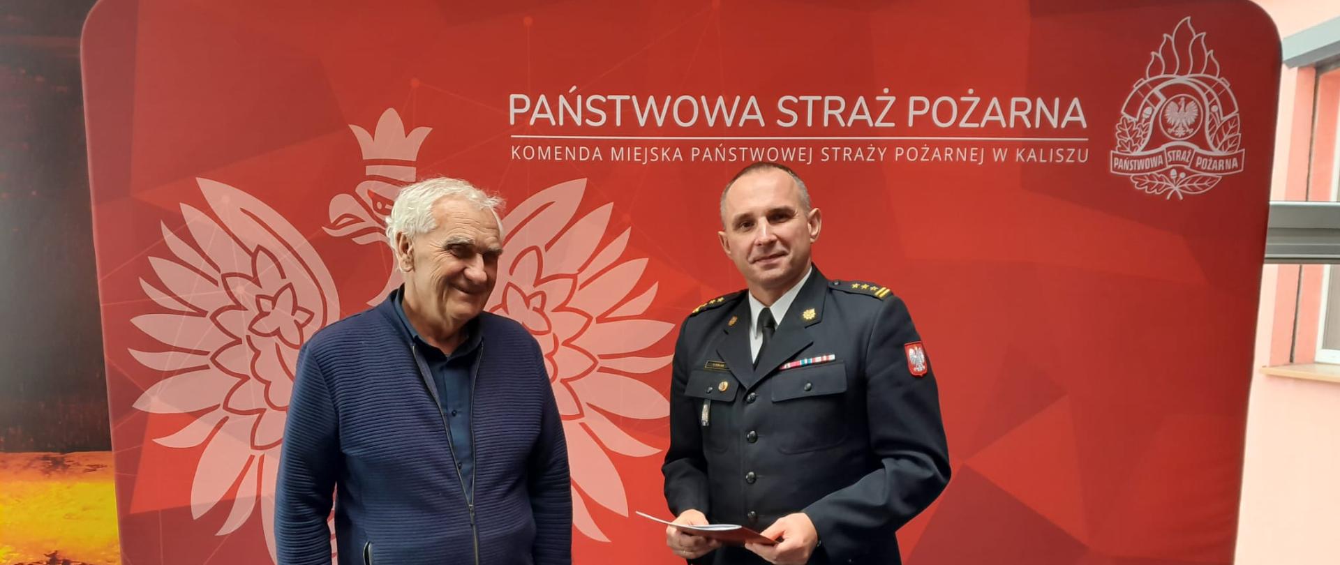 Na zdjęciu widzimy dwóch mężczyzn. Mężczyzna z prawej strony kadru to strażak, ubrany w mundur galowy kolor czarnego. Mężczyzna z lewej to cywil ubrany w granatową bluzę oraz oraz spodnie. Mężczyźni w dłoniach trzymają dokumenty. W tle widzimy czerwony baner koloru czerwonego z godłem polski oraz napisem Państwowa Straż Pożarna. 
