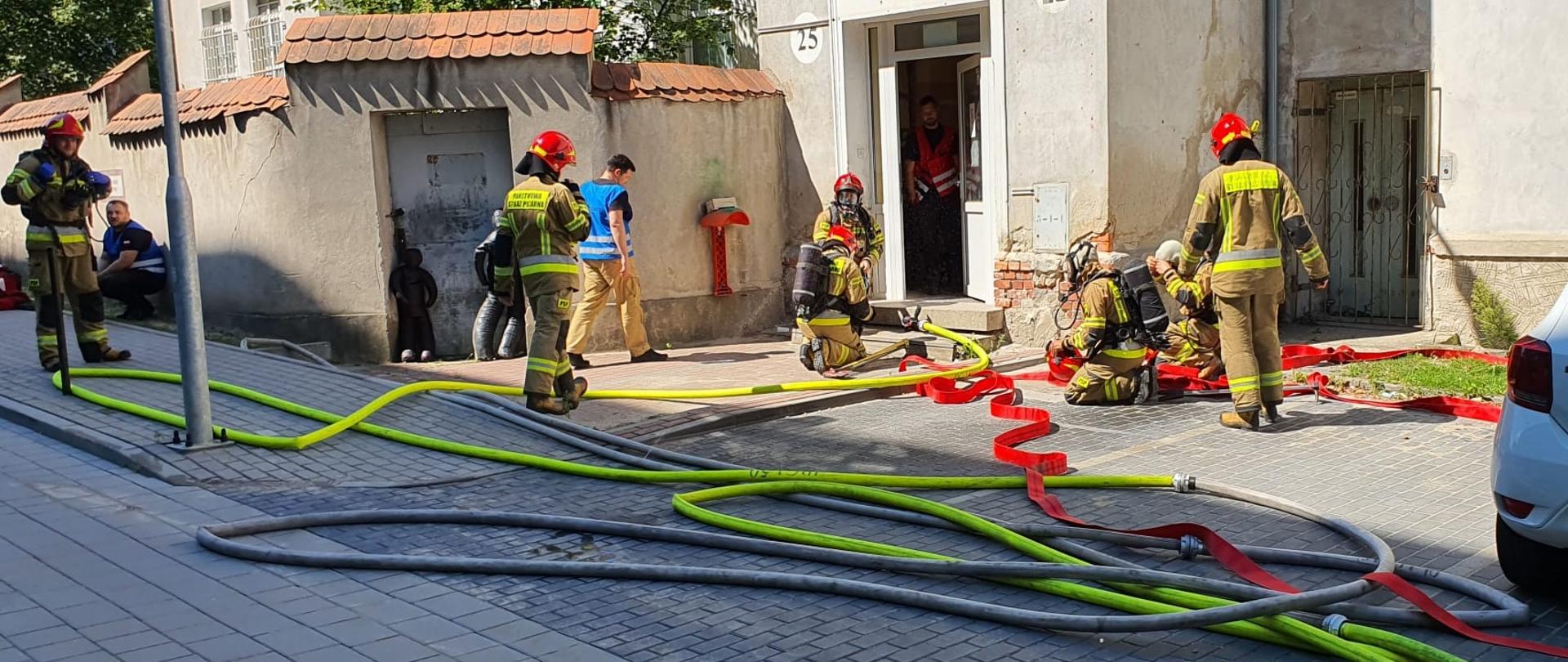 Strażacy przygotowują się do wejścia do zadymionej piwnicy - rozwijają linie gaśnicze i przygotowują sprzęt.