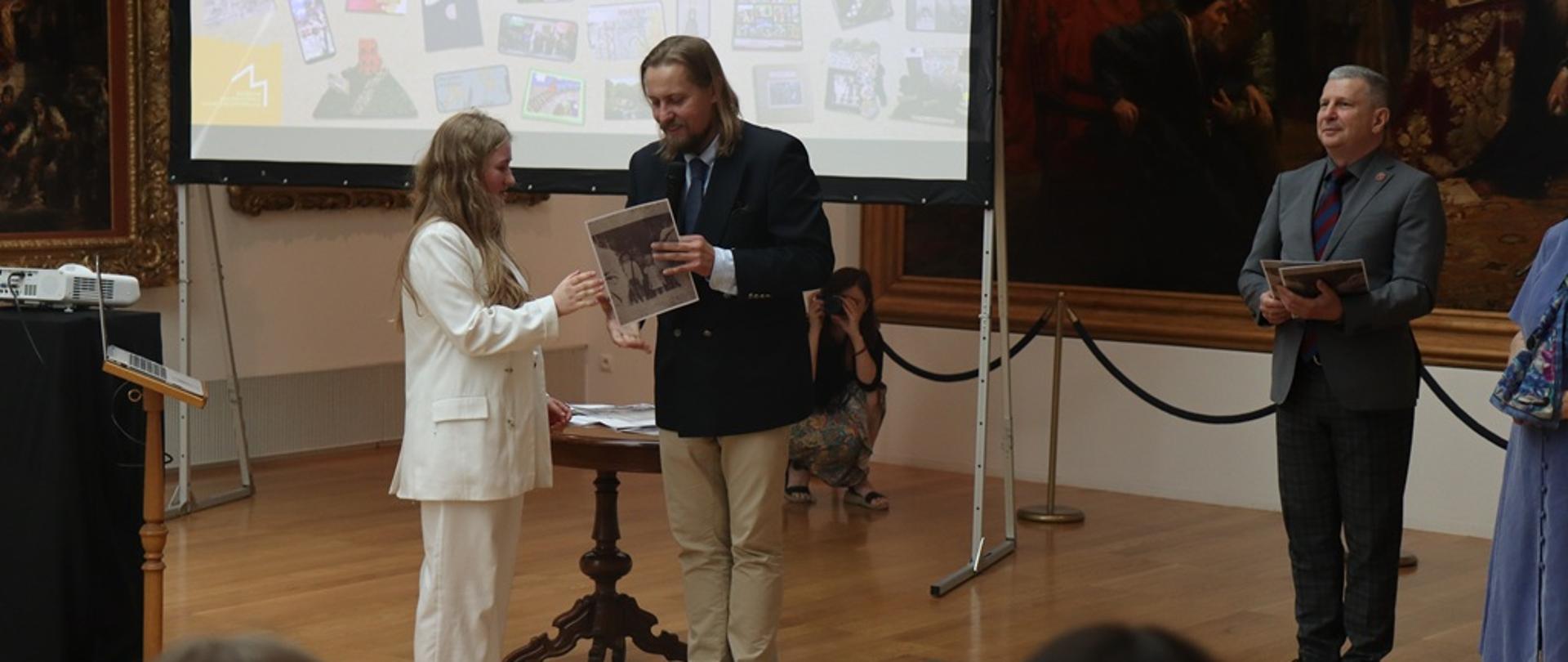 Scena z gali wręczenia nagród: mężczyzna w marynarce przekazuje czarno-białą fotografię młodej dziewczynie w białym stroju, obok stoi inny mężczyzna w garniturze, a w tle widać ekran z prezentacją prac.
