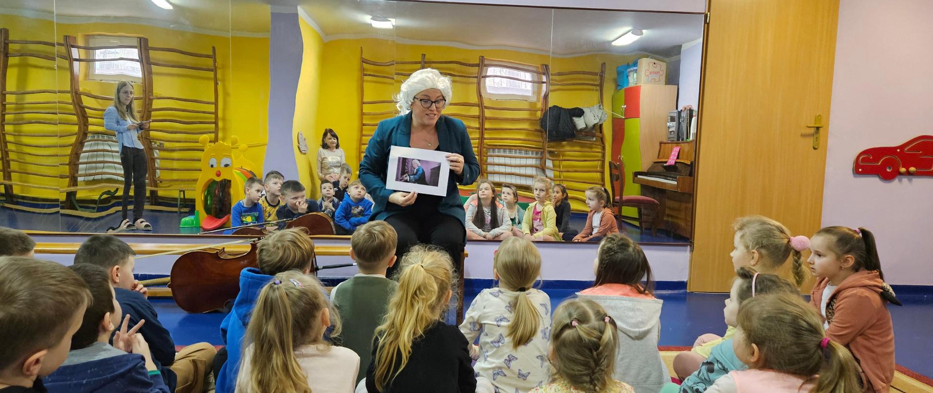 Zdjęcie przedstawia dzieci przysłuchujące się opowieści o Janie Sebastianie Bachu przedstawianej przez Panią Urszulę Właźniak. Pani Urszula trzyma w dłoniach grafikę przedstawiającą sylwetkę J. S.Bacha. Na głowie ma perukę barokową w kolorze białym. W tle lustra w których widać sylwetki dzieci.
