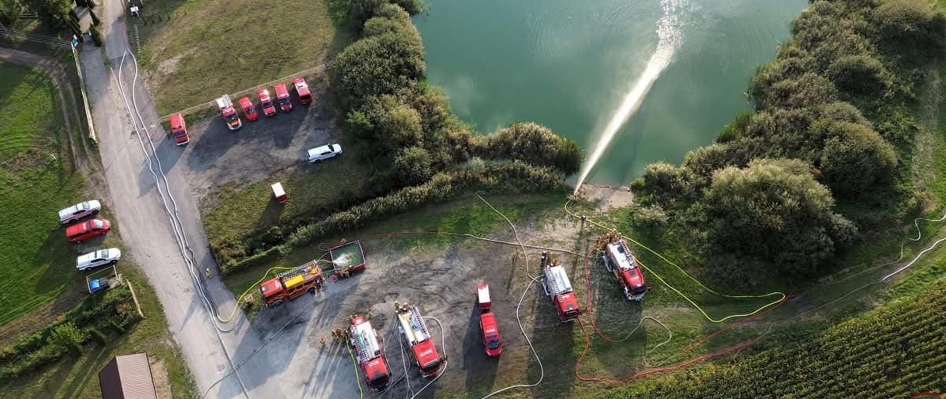 Widok z góry na jezioro, u brzegu którego ustawiono pojazdy gaśnicze. Z jednej linii gaśniczej woda podawana jest do jeziora. Widać drogę dojazdową, na skraju pole uprawne.