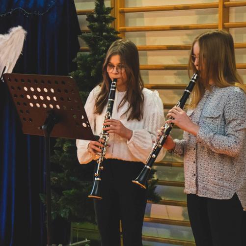 Zdjęcie przedstawia duet klarnetowy złożony z dziewczyn. Są one w trakcie wykonywania utworu muzycznego. Przed nimi stoi wysoki czarny pulpit z nutami. W tle widnieją drabinki gimnastyczne, choinka oraz niebieska kotara ze skrzydłami anioła jako dekoracja i napis "Hej kolęda"