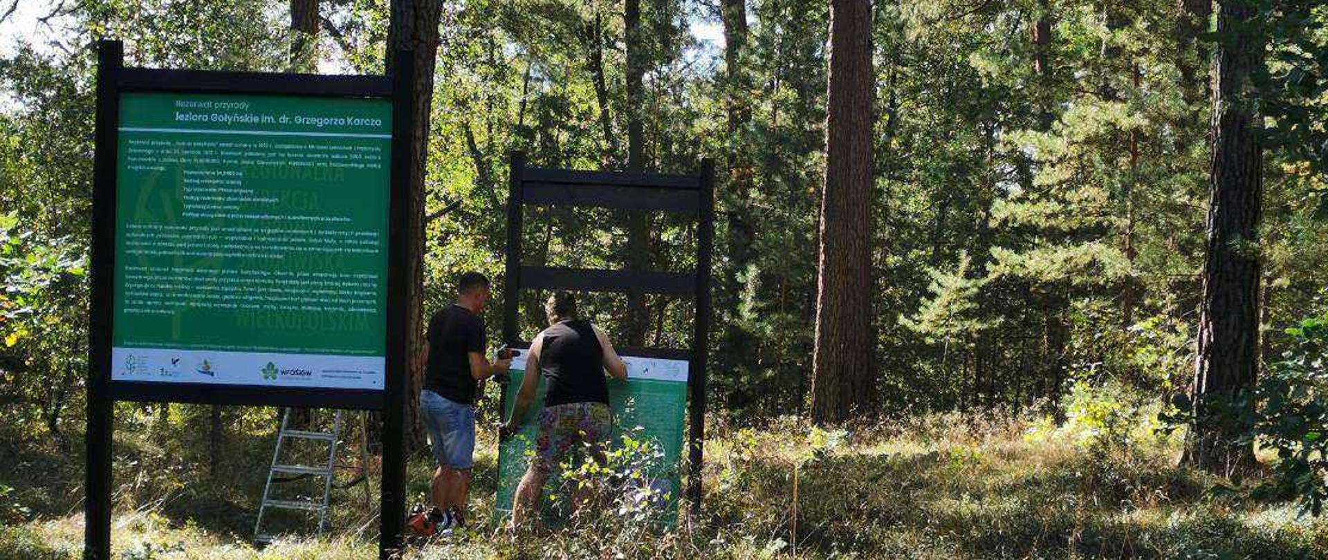 Na pierwszym planie tablica edukacyjna. W tle dwóch mężczyzn montujących nową tablicę.