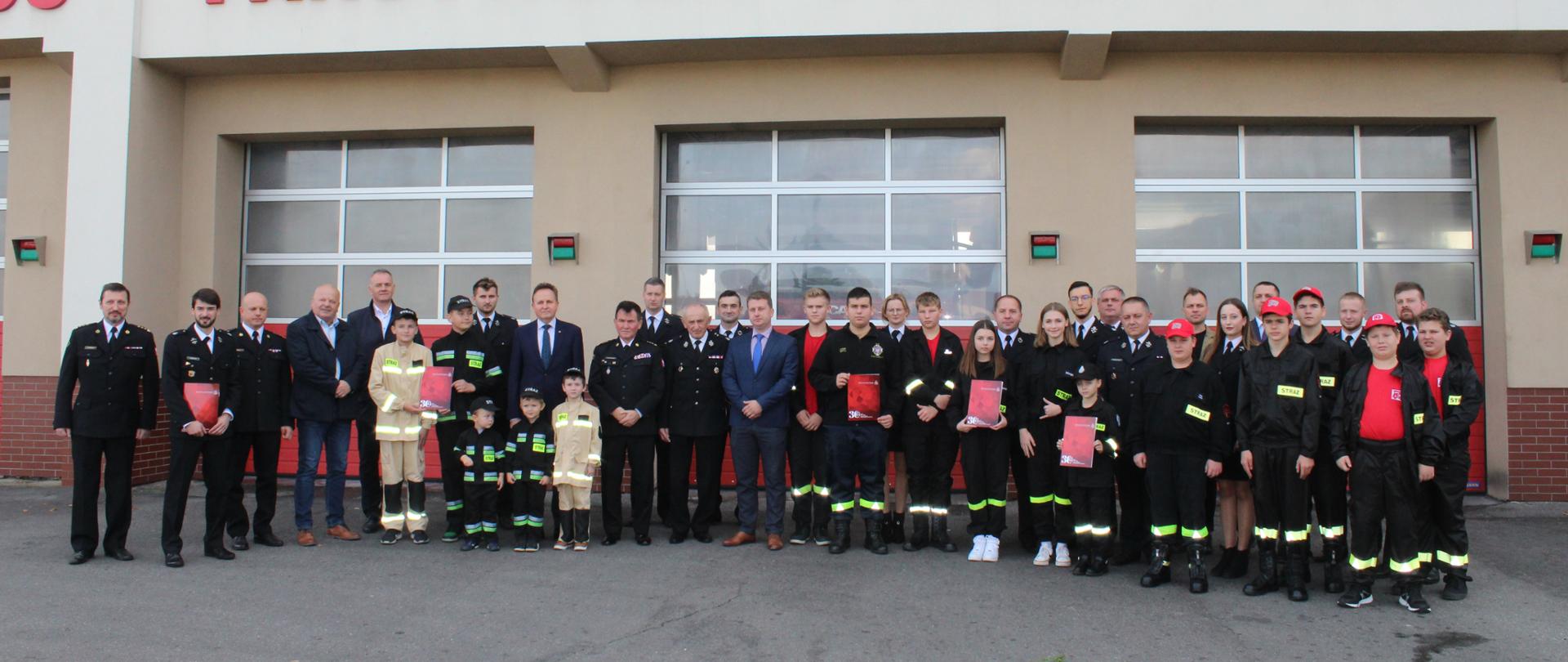 Członkowie młodzieżowych drużyn pożarniczych wraz ze swoimi opiekunami ubrani w mundury galowe, a także zaproszenie goście stoją przed bramami garażowymi na placu wewnętrznym przed budynkiem komendy