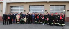 Członkowie młodzieżowych drużyn pożarniczych wraz ze swoimi opiekunami ubrani w mundury galowe, a także zaproszenie goście stoją przed bramami garażowymi na placu wewnętrznym przed budynkiem komendy
