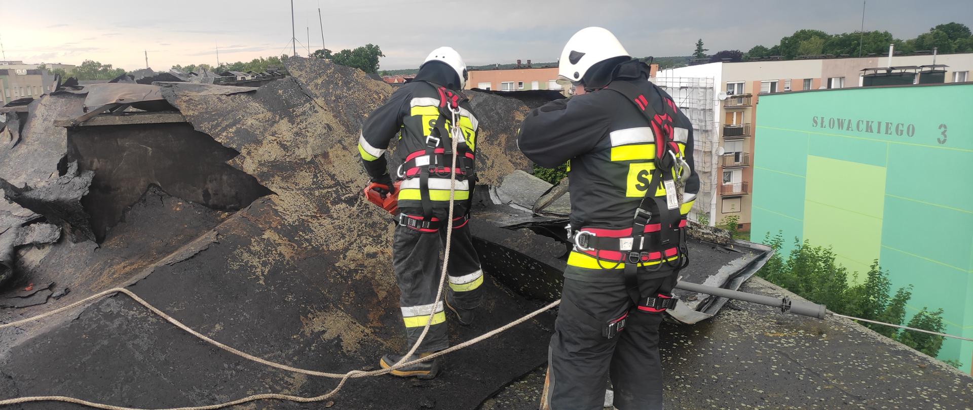 Na zdjęciu dwóch strażaków w ubraniach specjalnych i białych hełmach, zabezpieczeni linkami. Strażacy znajdują się na dachu usuwają zerwane pokrycie dachu.