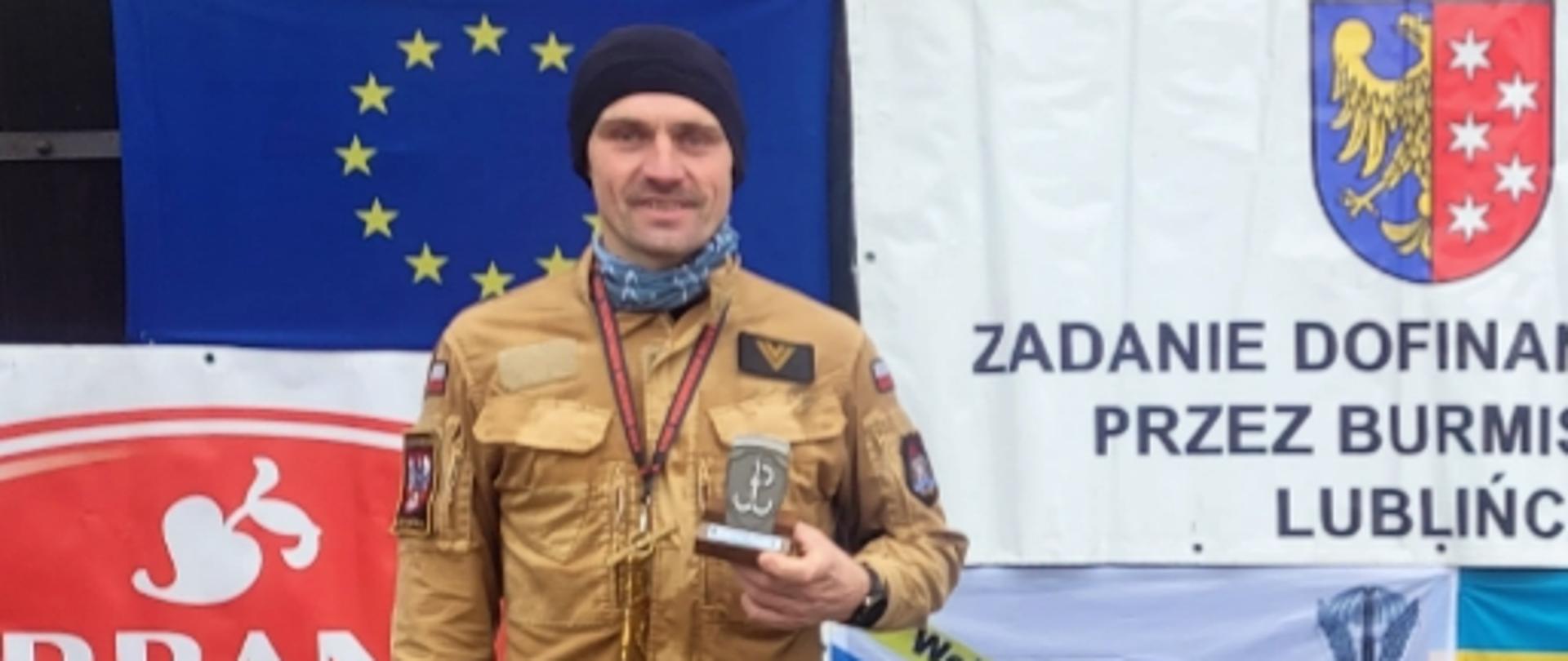 Dwóch mężczyzn w beżowych mundurach stoi na drewnianym podium podczas zimowej imprezy sportowej. Każdy trzyma w dłoni statuetkę oraz ma na szyi medal. W tle widoczne są liczne banery i flagi, m.in. z herbem miasta oraz napisami organizatora wydarzenia. Na ziemi leży śnieg, a wokół podium ustawione są nagrody.