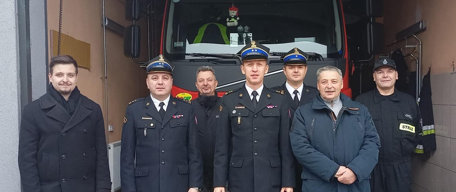 kilku strażaków oraz mężczyzn w ubraniach cywilnych stoi przed samochodem strażackim, samochód jest w garażu