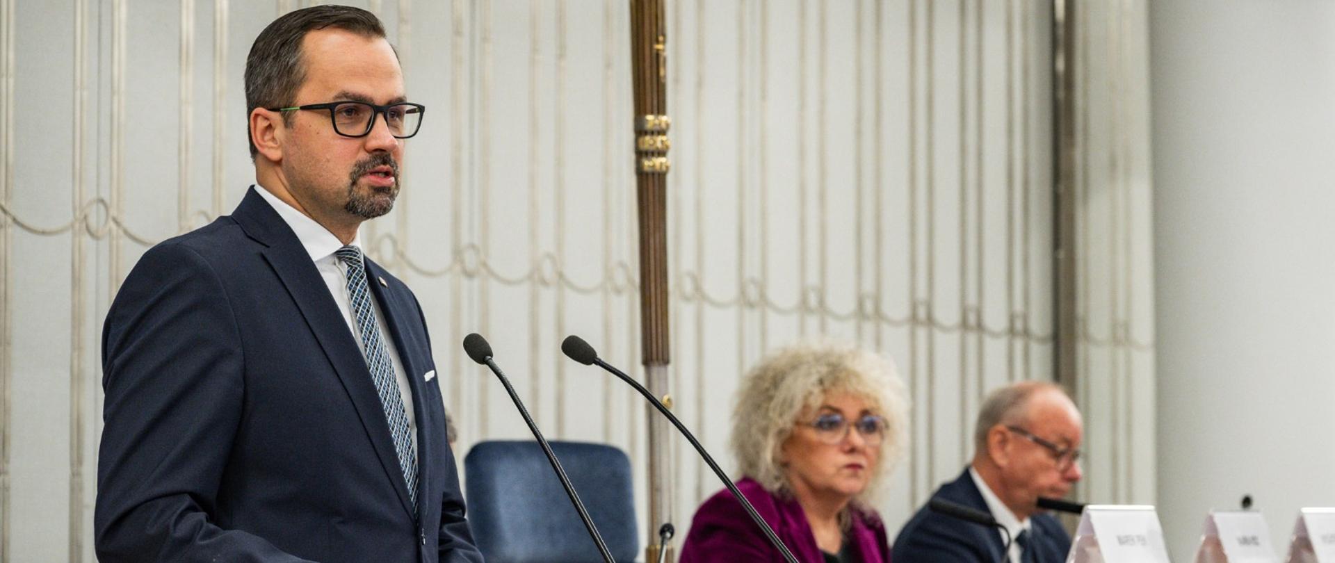 Wiceminister Marcin Horała stoi przy pulpicie i mówi do mikrofonu. Obok niego, po prawej stronie, siedzi wicemarszałek Senatu Maria koc oraz senator PiS.