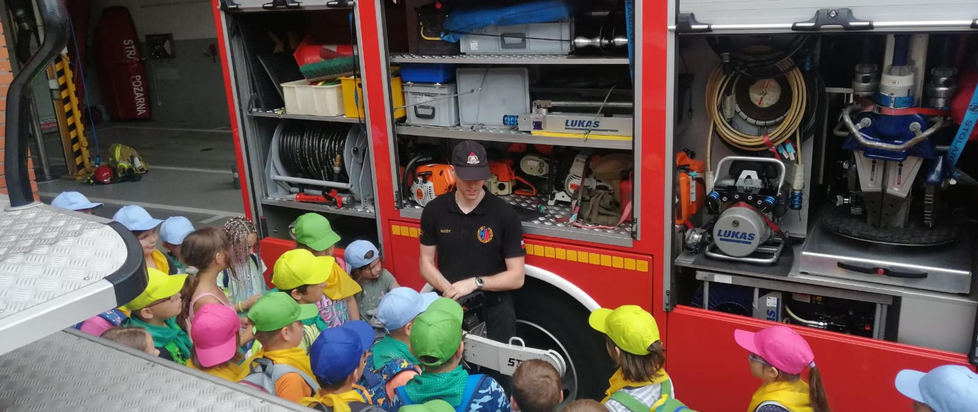 Zdjęcie przedstawia strażaka, którzy edukuje dzieci i młodzież.
W tle pojazd gaśniczy i budynek Komendy.
