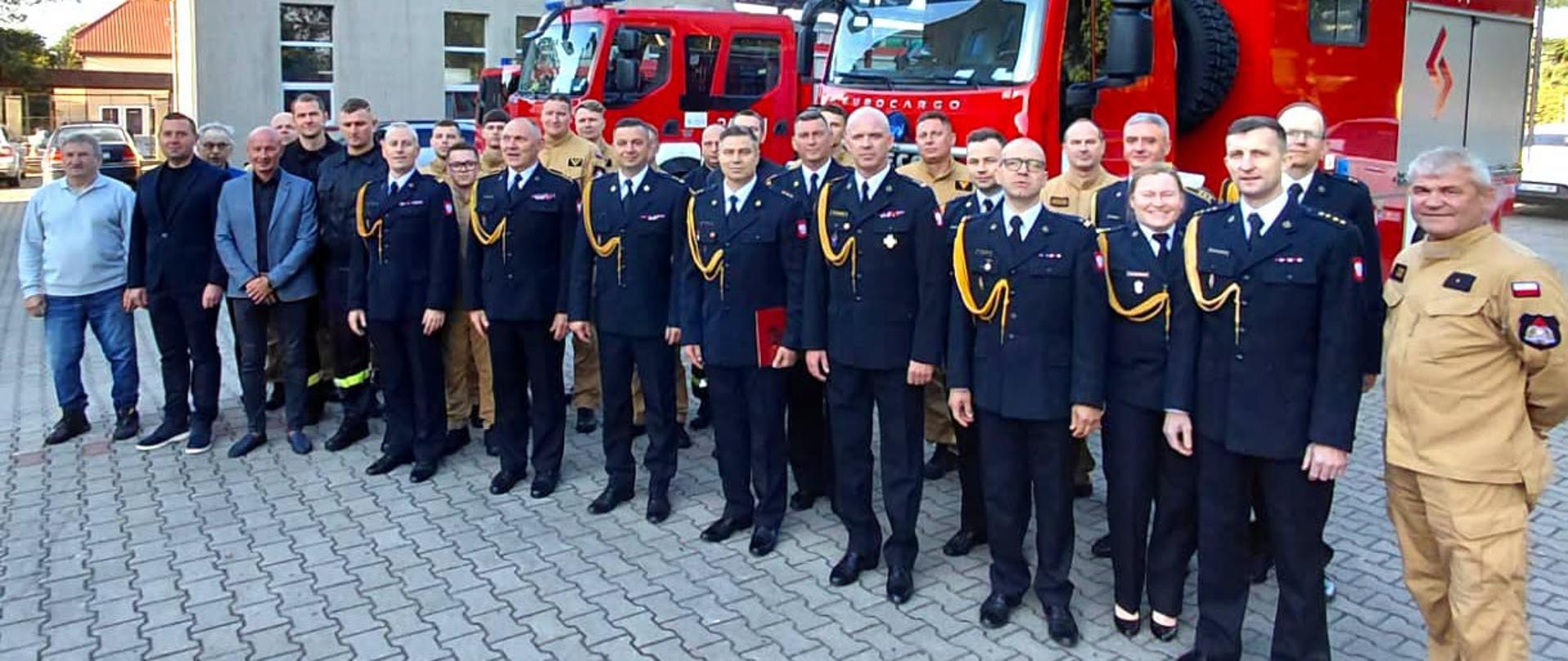 Zdjęcie przedstawia funkcjonariuszy PSP ubabrany w mundury galowe, oraz osoby cywilne podczas przekazywania obowiązków. w tle wozy strażackie. 