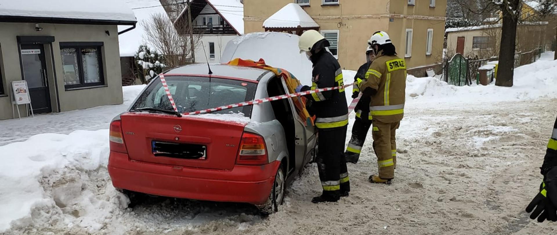 Po lewej stronie stojący na zaśnieżonym poboczu uszkodzony samochód. Przy nim stoi trzech strażaków w ubraniach specjalnych. W tle budynki.