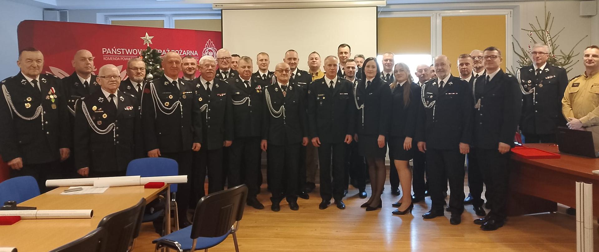 Grupowe zdjęcie przedstawiające strażaków Państwowej Straży Pożarnej oraz zaproszonych gości w galowych mundurach, ustawionych w sali konferencyjnej Komendy Powiatowej PSP. Uczestnicy stoją w kilku rzędach, pozując do wspólnej fotografii podczas uroczystości jubileuszu 30-lecia Krajowego Systemu Ratowniczo-Gaśniczego. W tle widoczny jest baner Państwowej Straży Pożarnej, ekran projekcyjny oraz elementy świątecznego wystroju.
