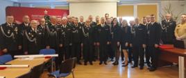 Grupowe zdjęcie przedstawiające strażaków Państwowej Straży Pożarnej oraz zaproszonych gości w galowych mundurach, ustawionych w sali konferencyjnej Komendy Powiatowej PSP. Uczestnicy stoją w kilku rzędach, pozując do wspólnej fotografii podczas uroczystości jubileuszu 30-lecia Krajowego Systemu Ratowniczo-Gaśniczego. W tle widoczny jest baner Państwowej Straży Pożarnej, ekran projekcyjny oraz elementy świątecznego wystroju.