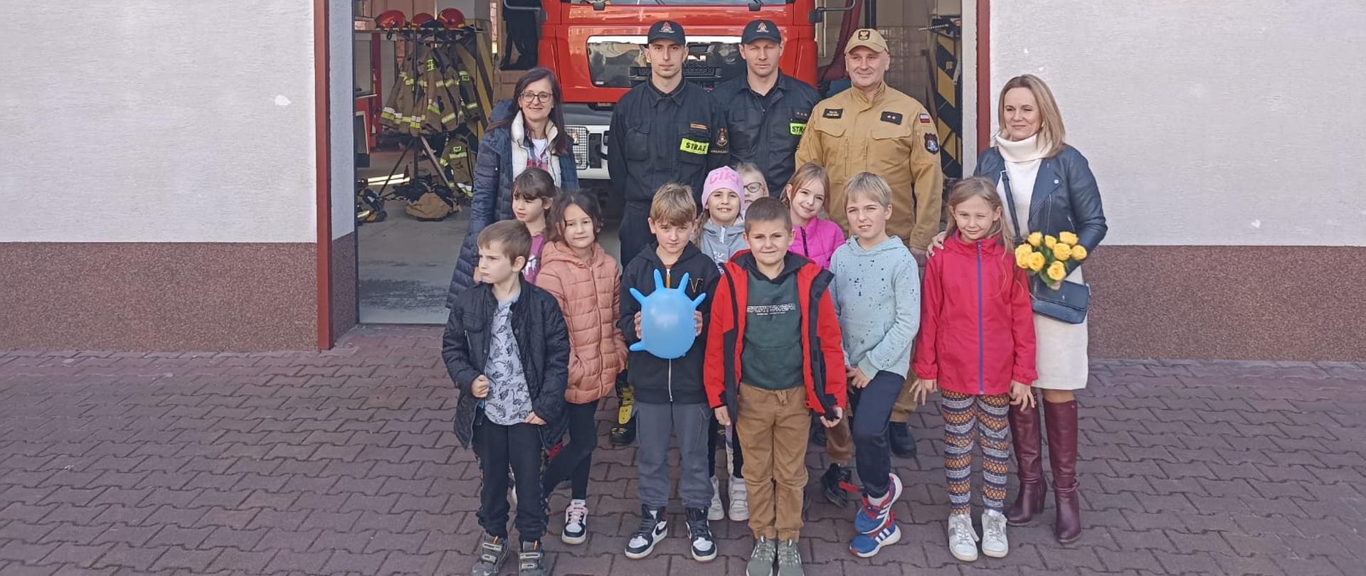 Zdjęcie przedstawia trzech strażaków, dwie nauczycielki oraz dzieci. Wszyscy stoją na tle garażu komendy w którym stoi samochód gaśniczy. Na ścianie wisi logo Państwowej Straży Pożarnej.