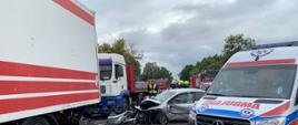 Zdjęcie przedstawia rozbity samochód osobowy marki Toyota Corolla na tle pojazdów strażackich oraz tirów.