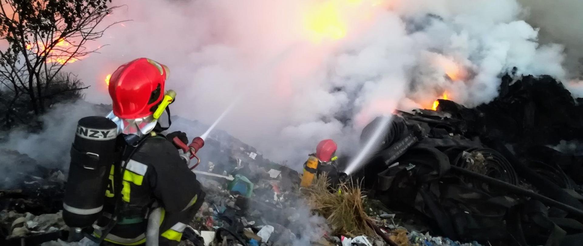 Dwóch strażaków w ubraniach specjalnych, chełmach oraz z butlami na plecach gaszący palącą się hałdę śmieci