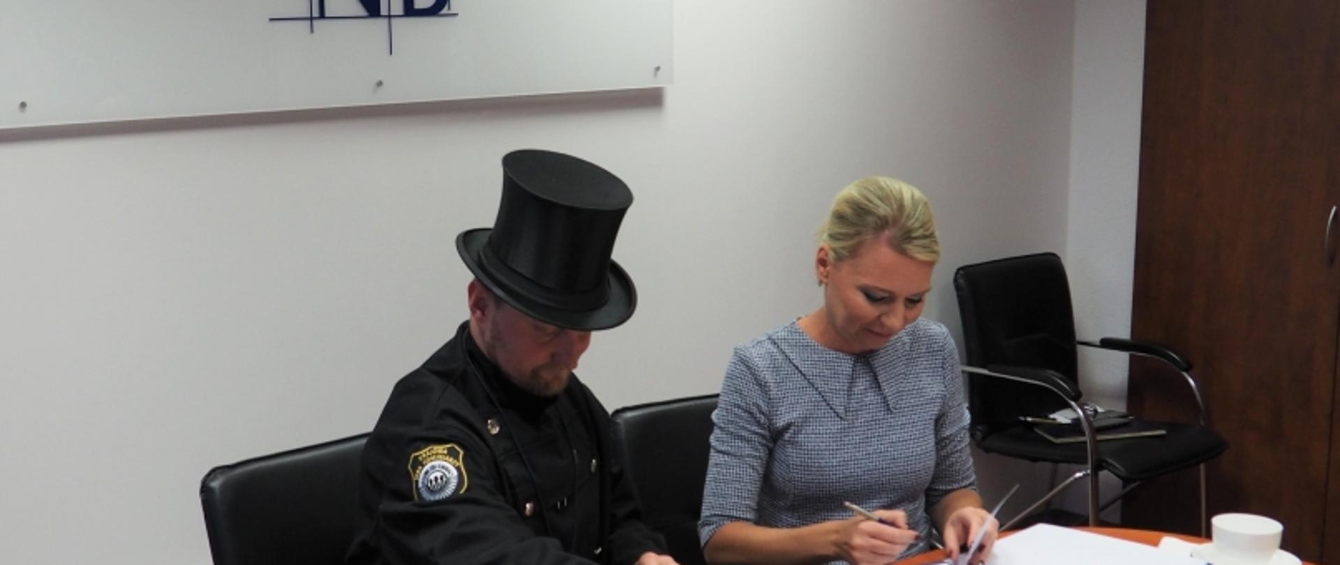 Dwie osoby przy stole z dokumentami; jedna w mundurze i cylindrze, druga w stroju biznesowym; w tle znak „KRD”.