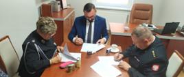 Fotografia przedstawia trzy osoby siedzące przy stole i podpisujące dokumenty.
