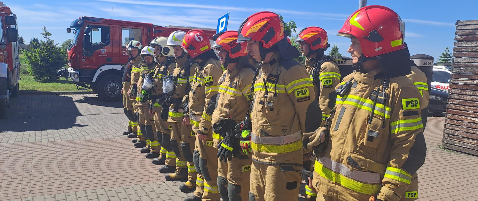 Zdjęcie przedstawia strażaków Państwowej Straży Pożarnej oraz strażaków Ochotniczych Straży Pożarnych ubranych w ubrania specjalne koloru piaskowego podczas odprawy przez ćwiczeniami.