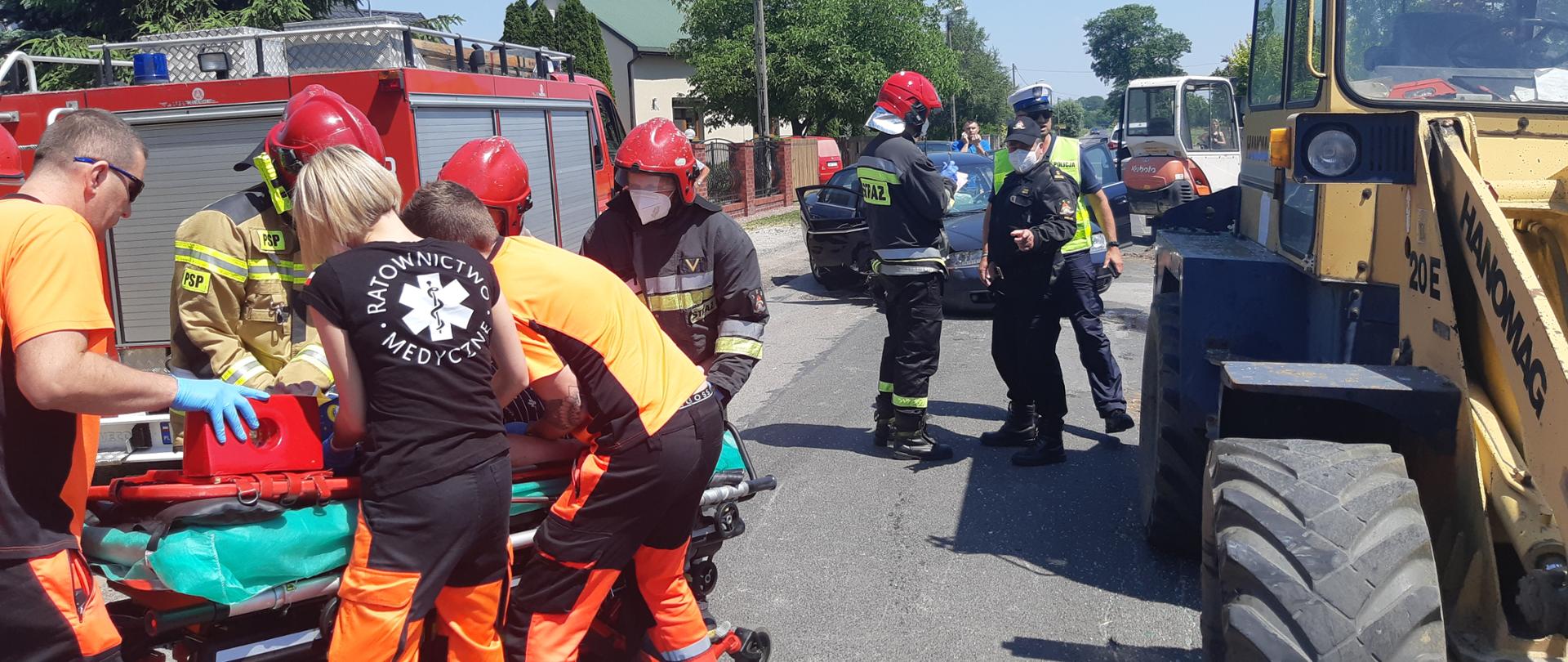 Zdjęcie przedstawia samochód osobowy po wypadku z ładowarką, strażaków i ratowników medycznych z osobą poszkodowaną, w tle samochód bojowy straży pożarnej.