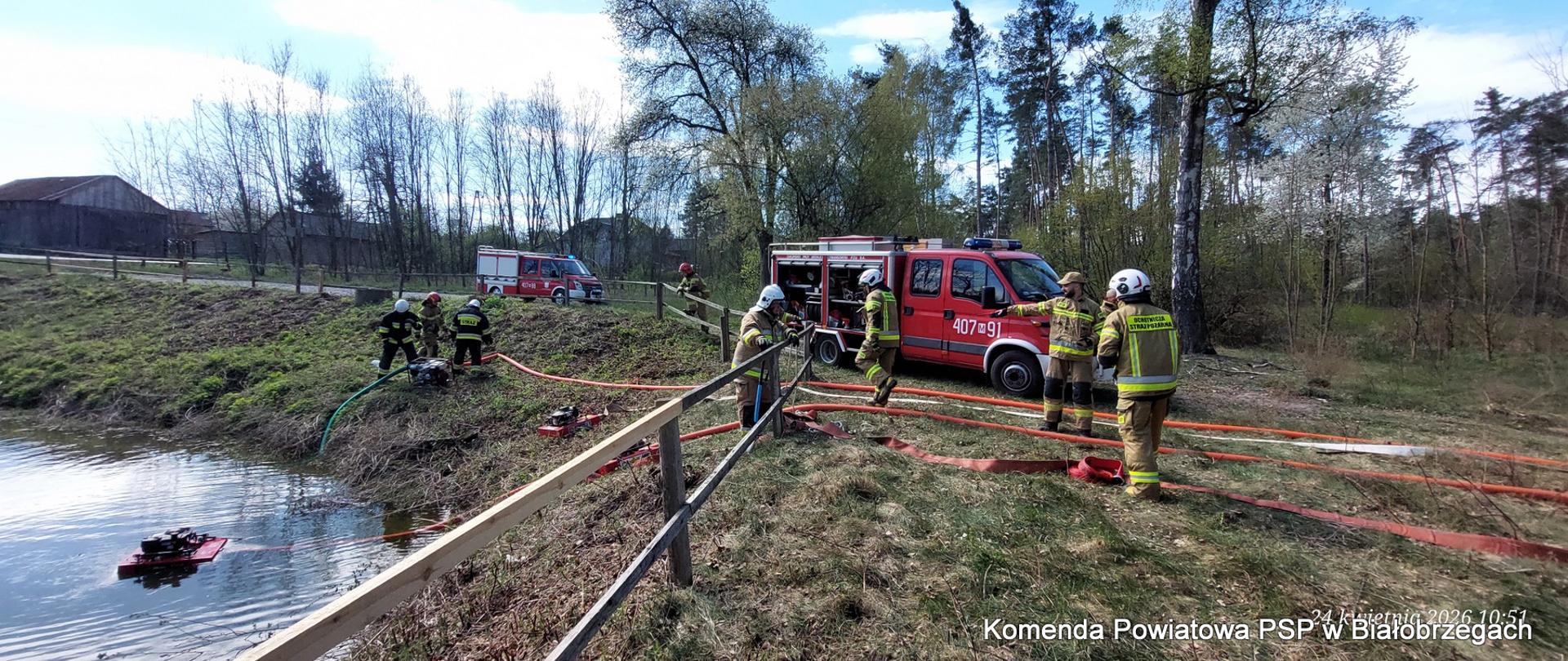 Akcja strażacka przy zbiorniku wodnym. Strażacy w piaskowych mundurach obsługują motopompy ustawione przy brzegu i na wodzie. Czerwony wóz techniczny stoi na skarpie, a od niego w stronę wody prowadzą liczne węże strażackie.